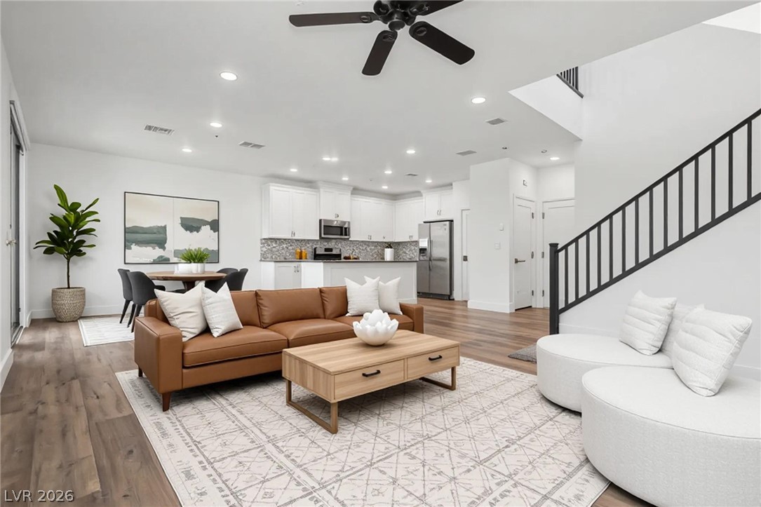Staged living room with brown sofa, white chairs, open kitchen, and black-railed staircase.