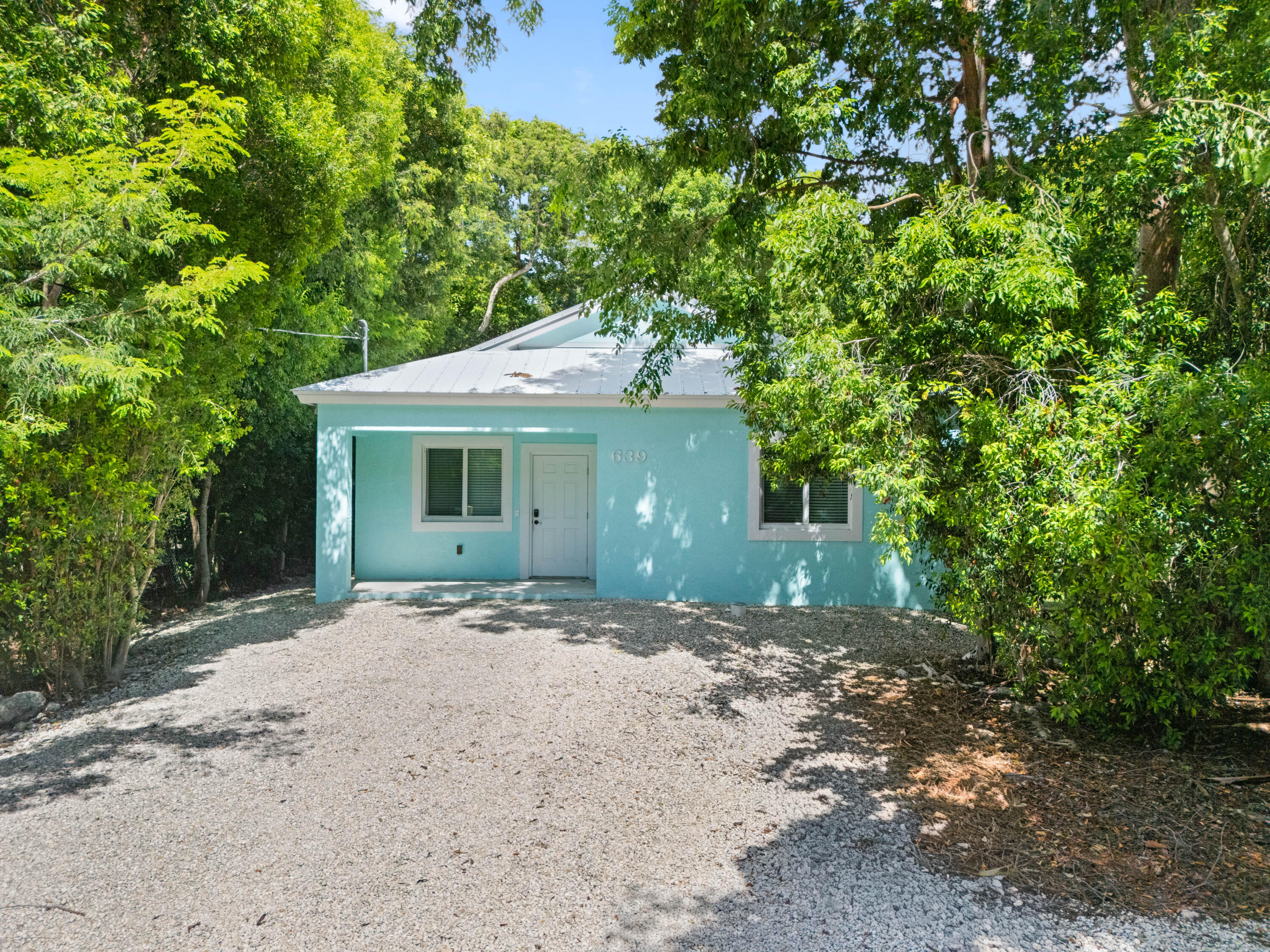 Turquoise cottage with a white metal roof, set among dense trees beside a gravel drive.