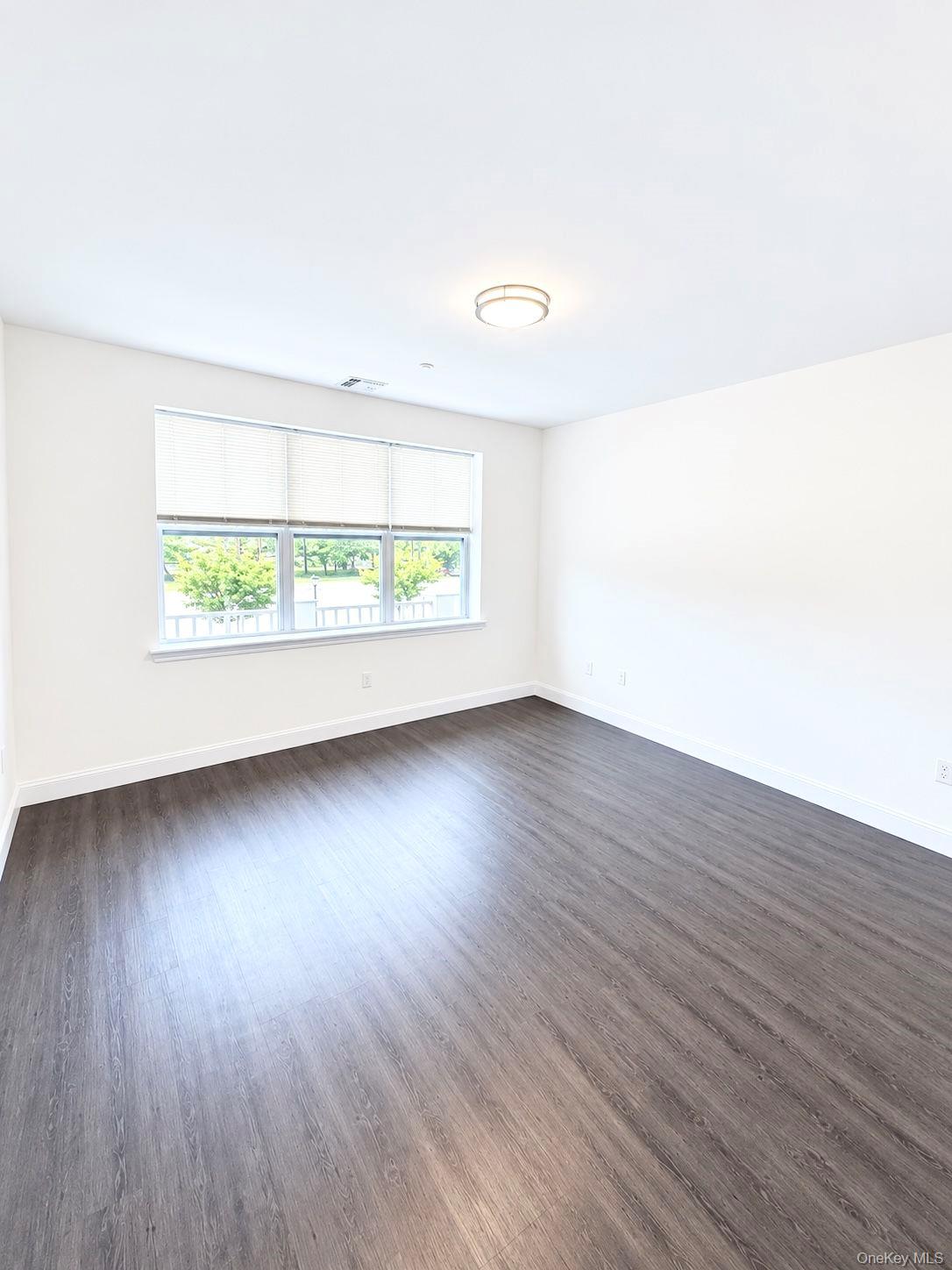 Empty room with dark plank flooring and a wide window with blinds overlooking greenery.
