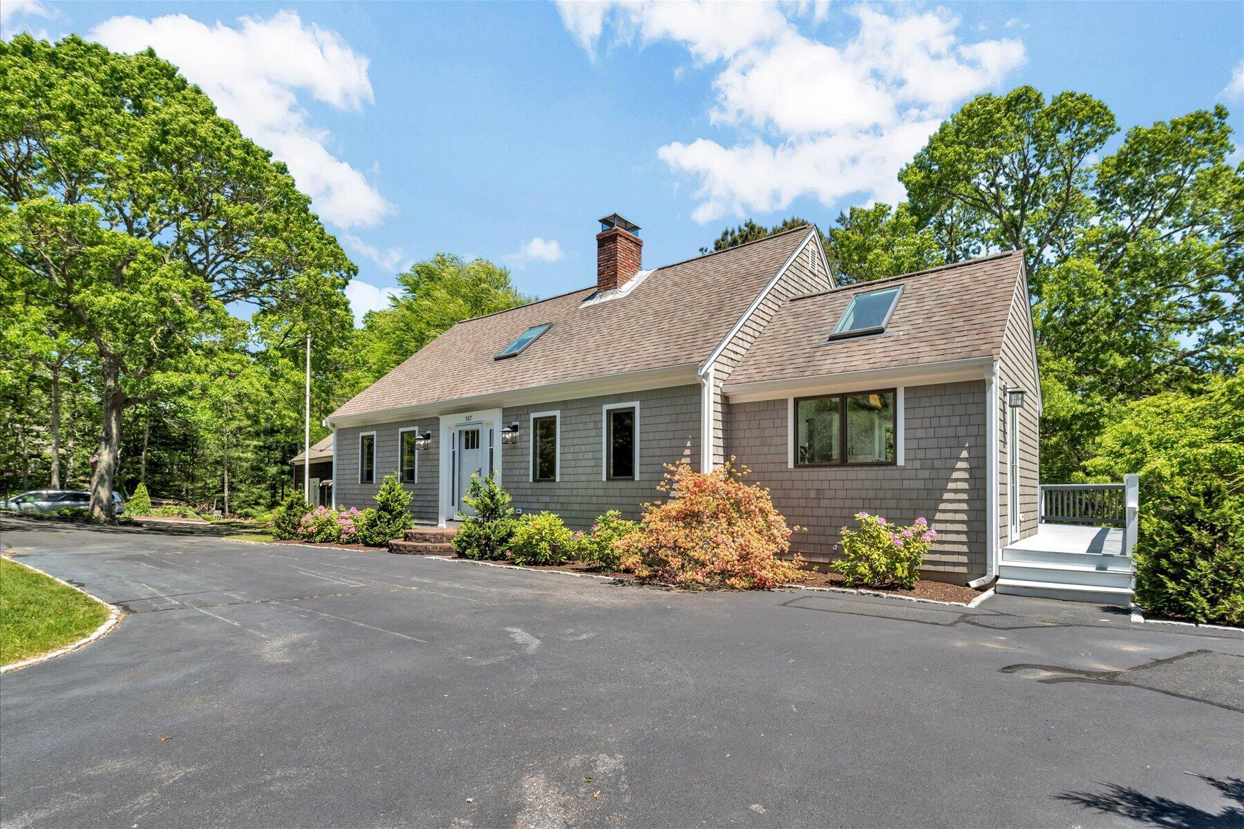 Gray shingle-sided home with landscaped flower beds, broad paved driveway, and a small white side deck.