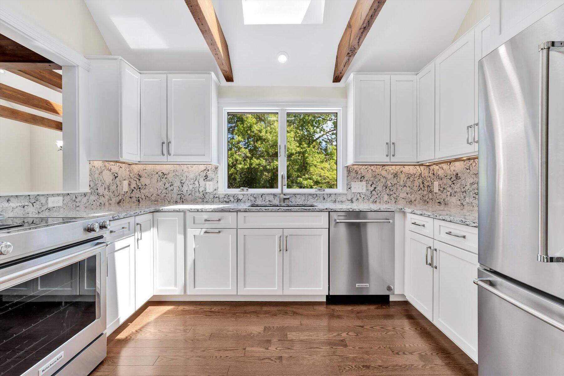 Bright kitchen with white cabinets, stone counters, stainless steel appliances, and exposed wood ceiling beams.