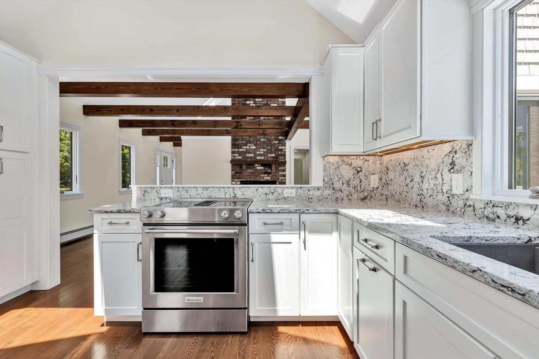 Kitchen with white cabinetry, veined stone counters, stainless range, and exposed beams leading to a brick fireplace.