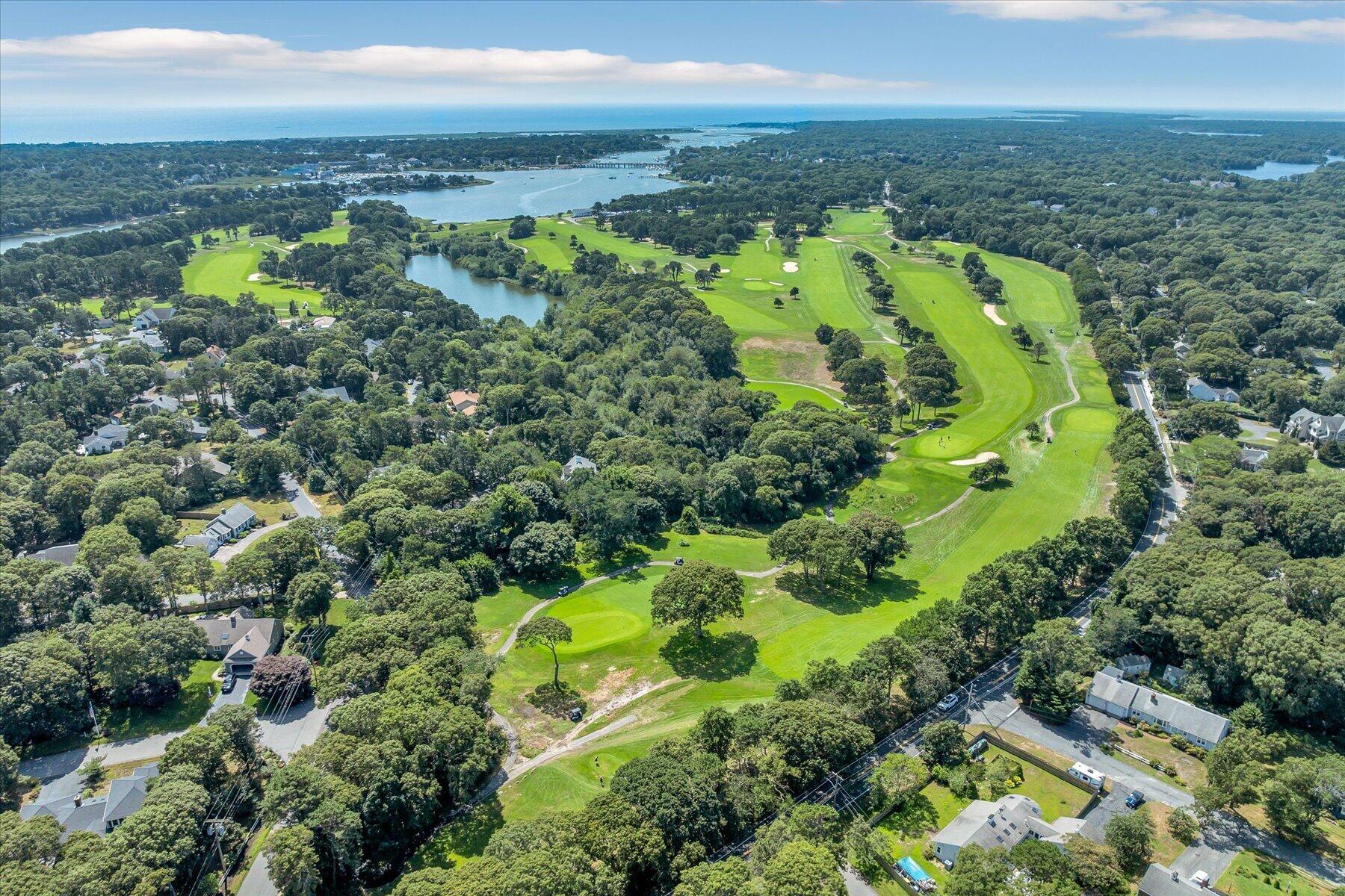 Aerial view of a golf course beside wooded neighborhoods and coastal water.
