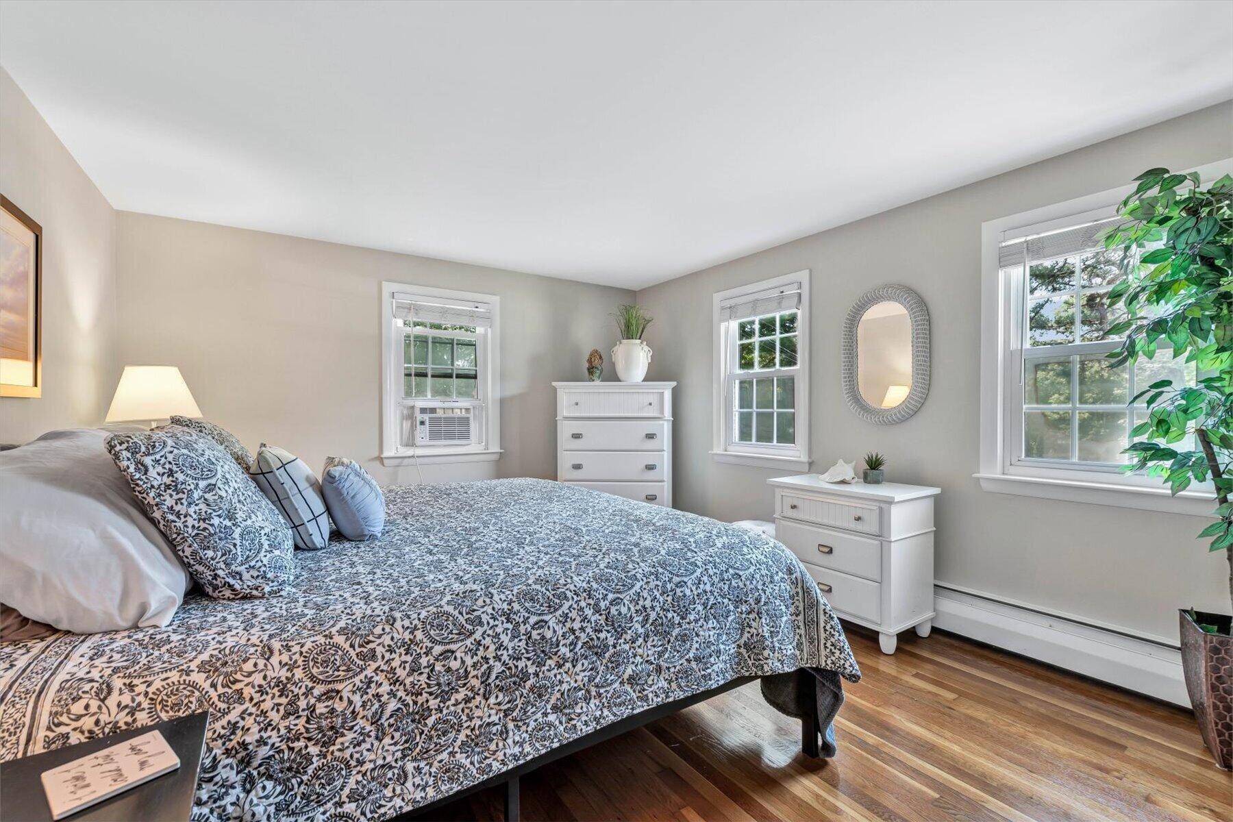 Bedroom with a patterned bedspread, white dressers, and three bright windows.