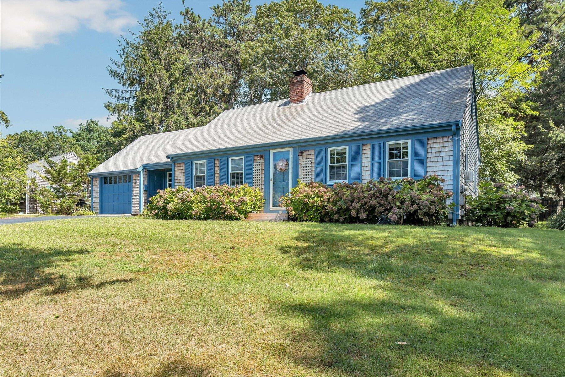 Shingle-sided Cape-style home with blue shutters, an attached garage, and flowering shrubs along the front.
