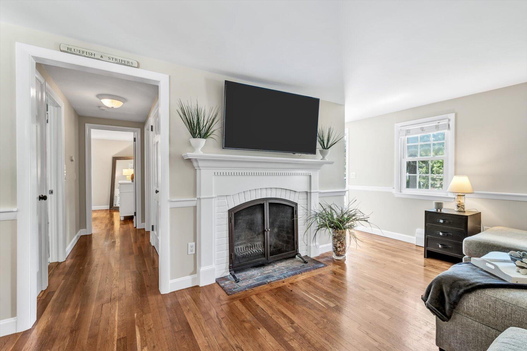 Living room with a white brick fireplace, mounted television, hardwood floors, and a hallway beyond.
