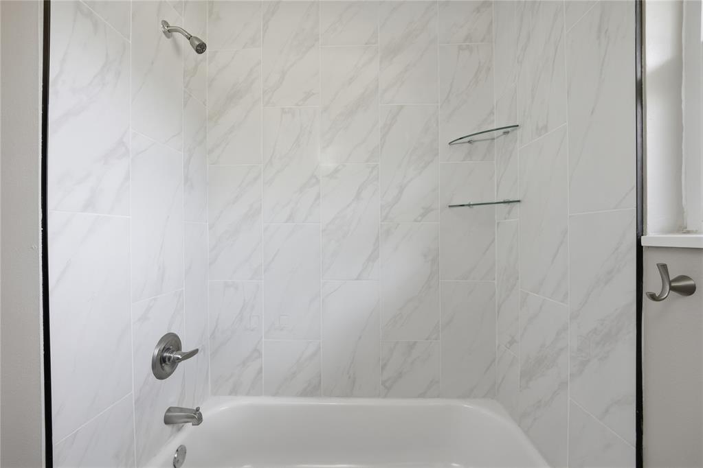 Bathtub with a white marble-look tile surround, chrome fixtures, and glass shelves.
