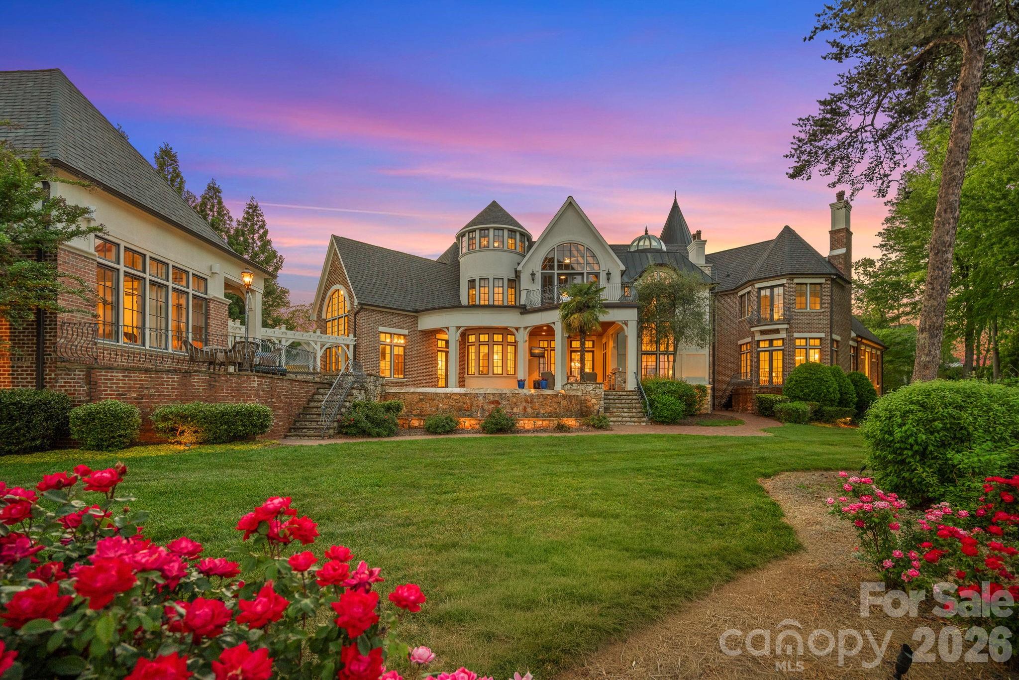 Large brick-and-stucco home with turret and arched windows, warmly lit at dusk behind a rose garden.