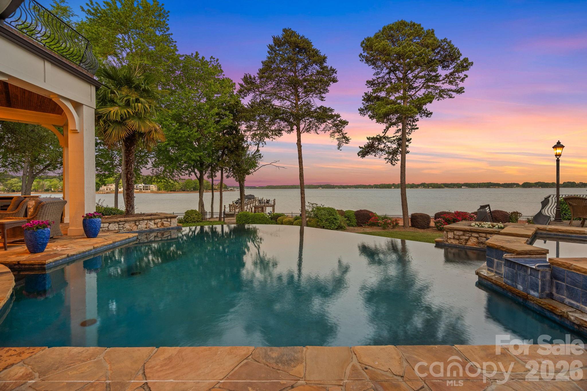 Lakefront pool and stone patio overlook the water at sunset, with a dock and covered seating area.