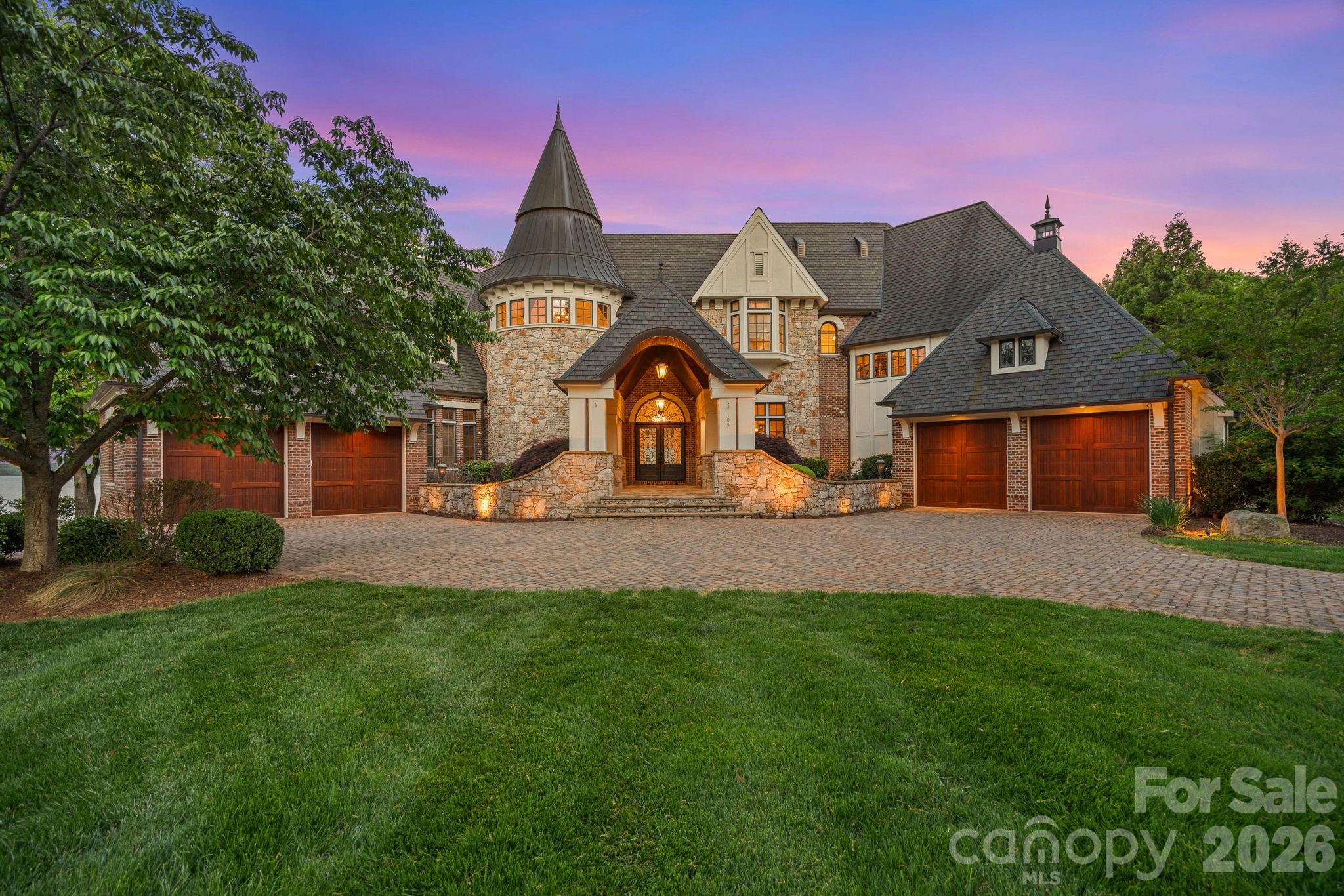 Luxury stone-and-brick home with a turret and multiple garages, illuminated at dusk.