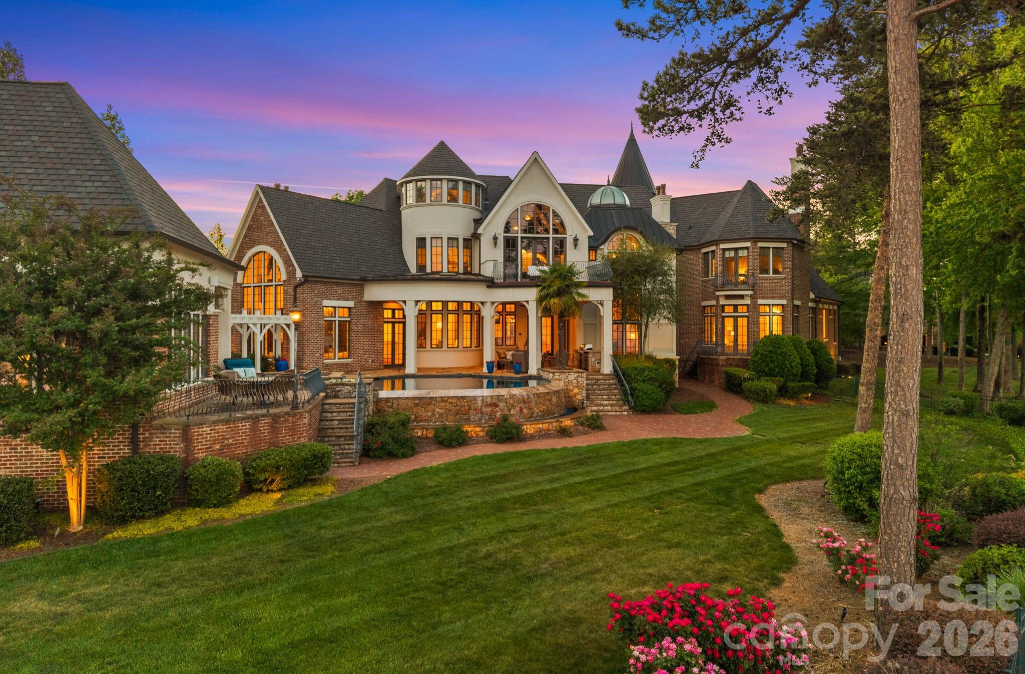 Large brick-and-stone home with turreted roof, illuminated windows, terrace pool, and landscaped lawn at dusk.