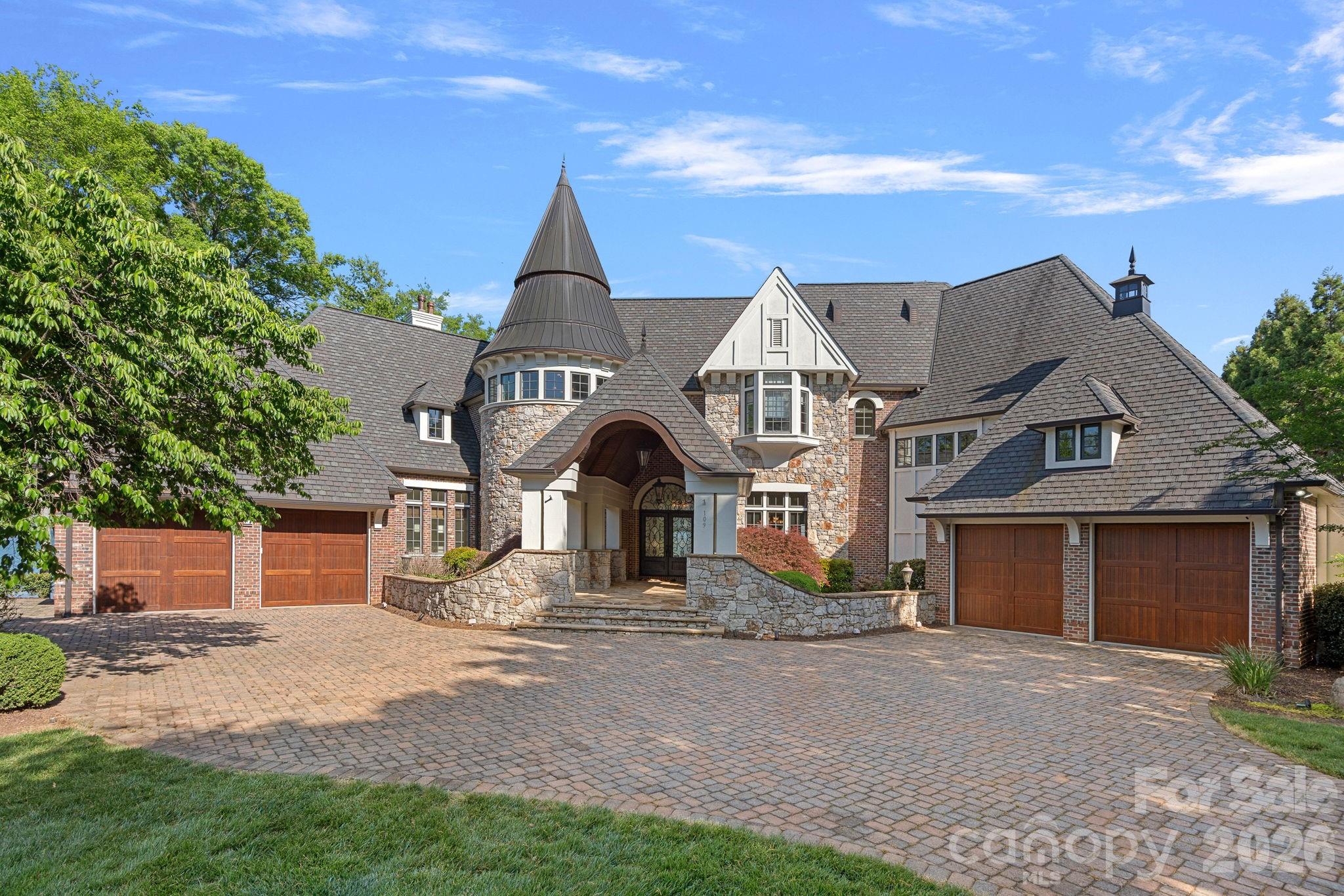 Stone-and-brick home with a round turret, arched entry, and broad paved driveway.