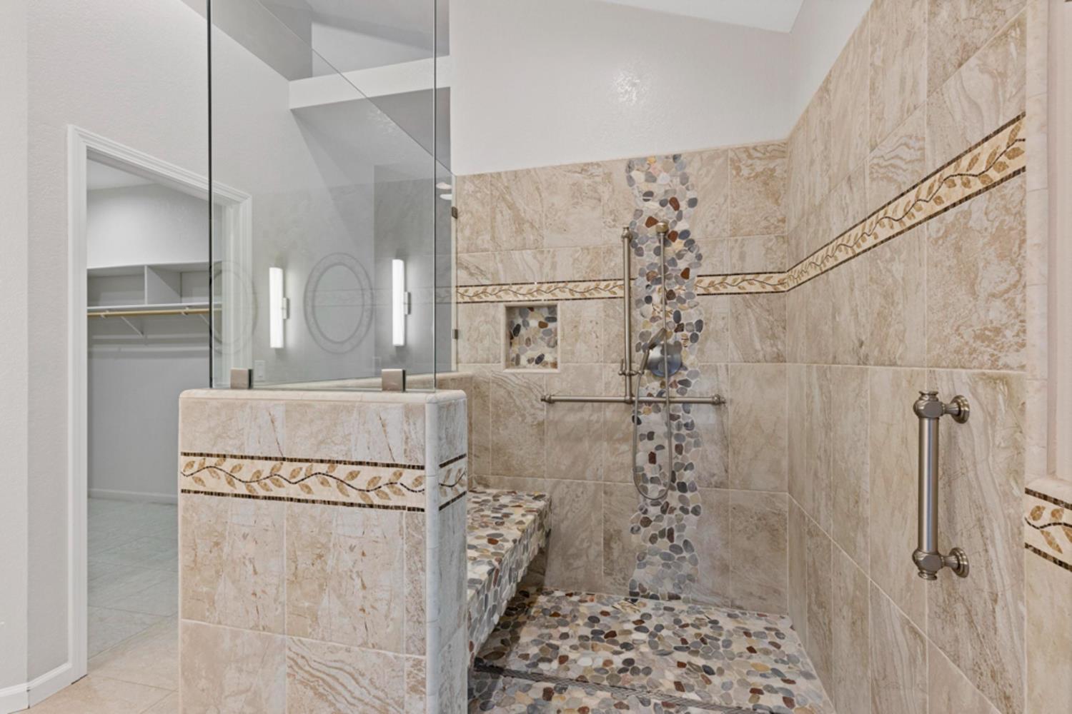 Walk-in shower with beige stone tile, pebble accents, a built-in bench, and grab bars.