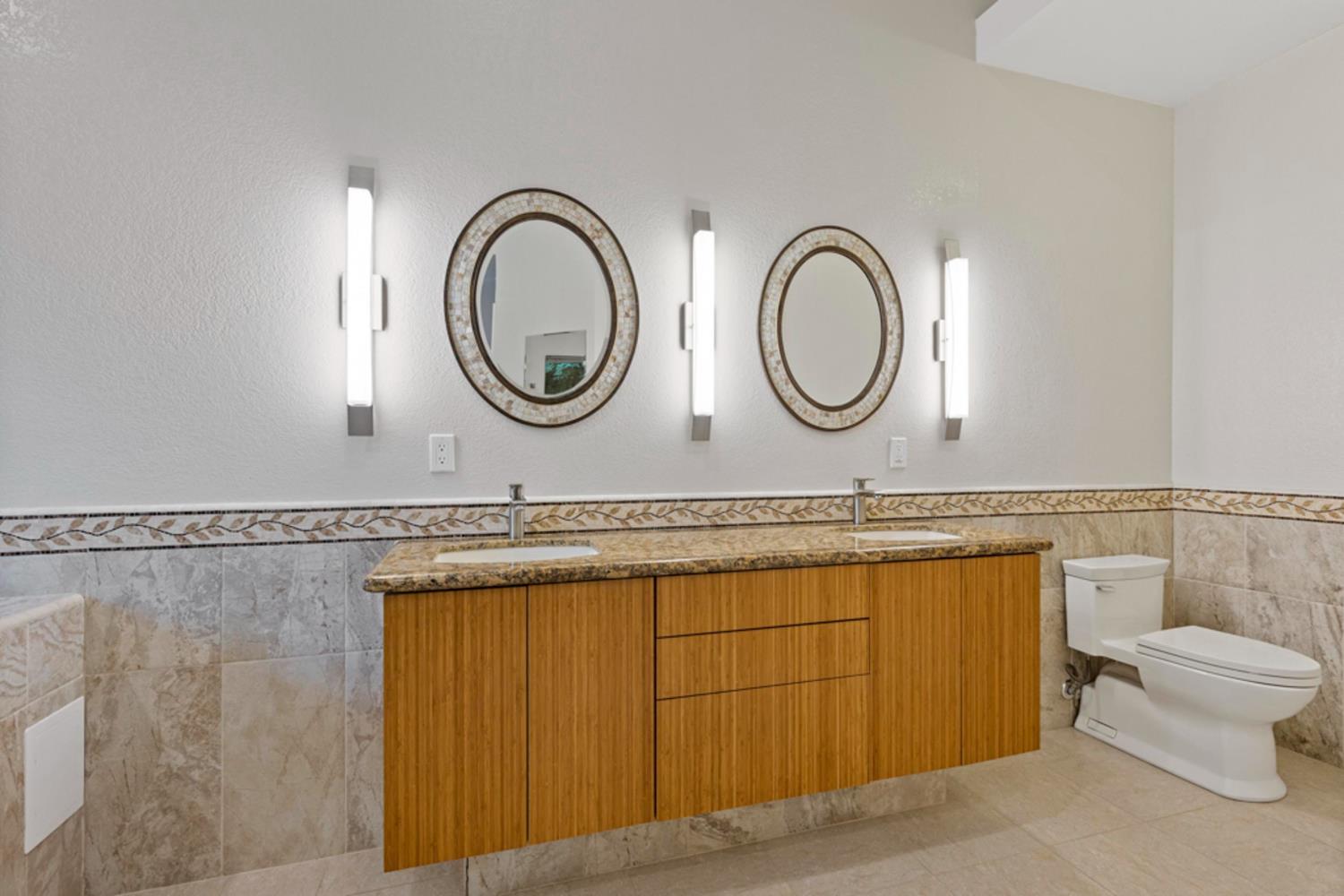 Bathroom with a double-sink wood vanity, patterned oval mirrors, tiled walls, and a toilet.