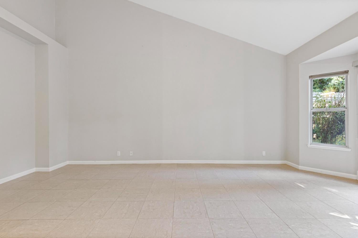 Empty living room with beige tile floors, high angled ceiling, and a window overlooking greenery.