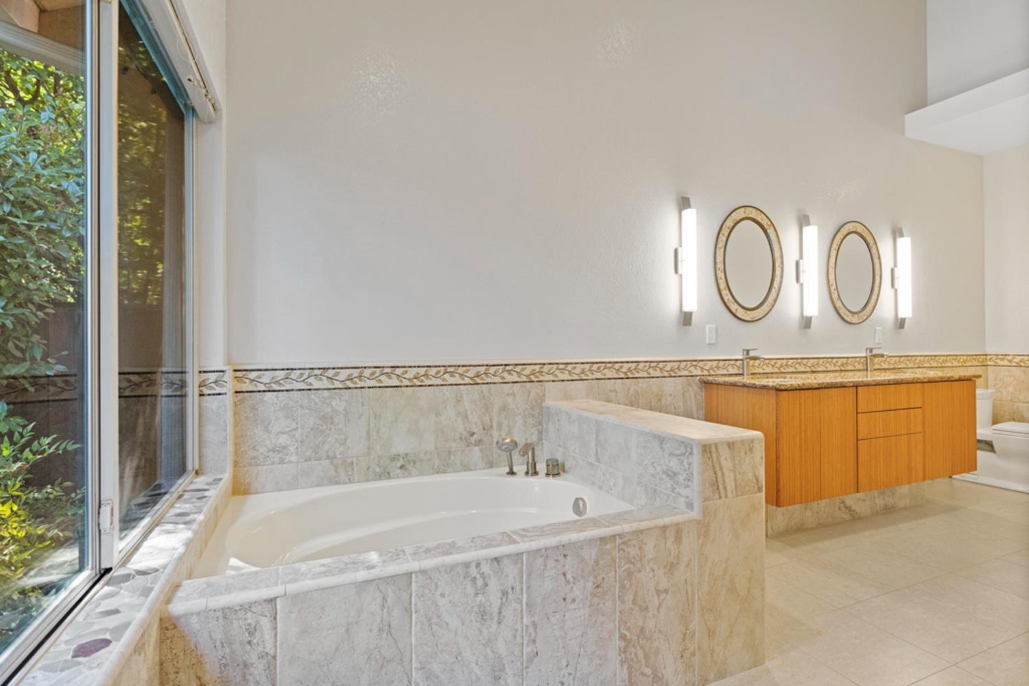 Primary bathroom with a marble-surround soaking tub beside a large window and a double-sink wood vanity.