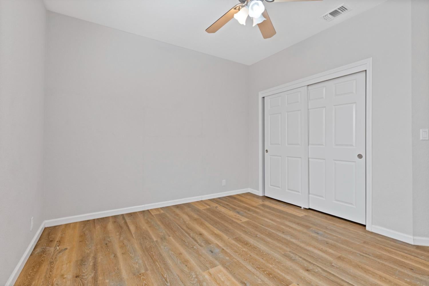 Empty bedroom with wood-look plank flooring, white sliding closet doors, and a ceiling fan.