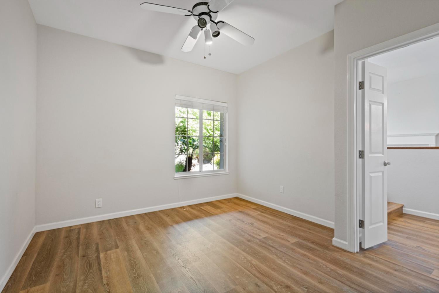 Empty room with wood-look floors, a ceiling fan, and a window overlooking greenery.