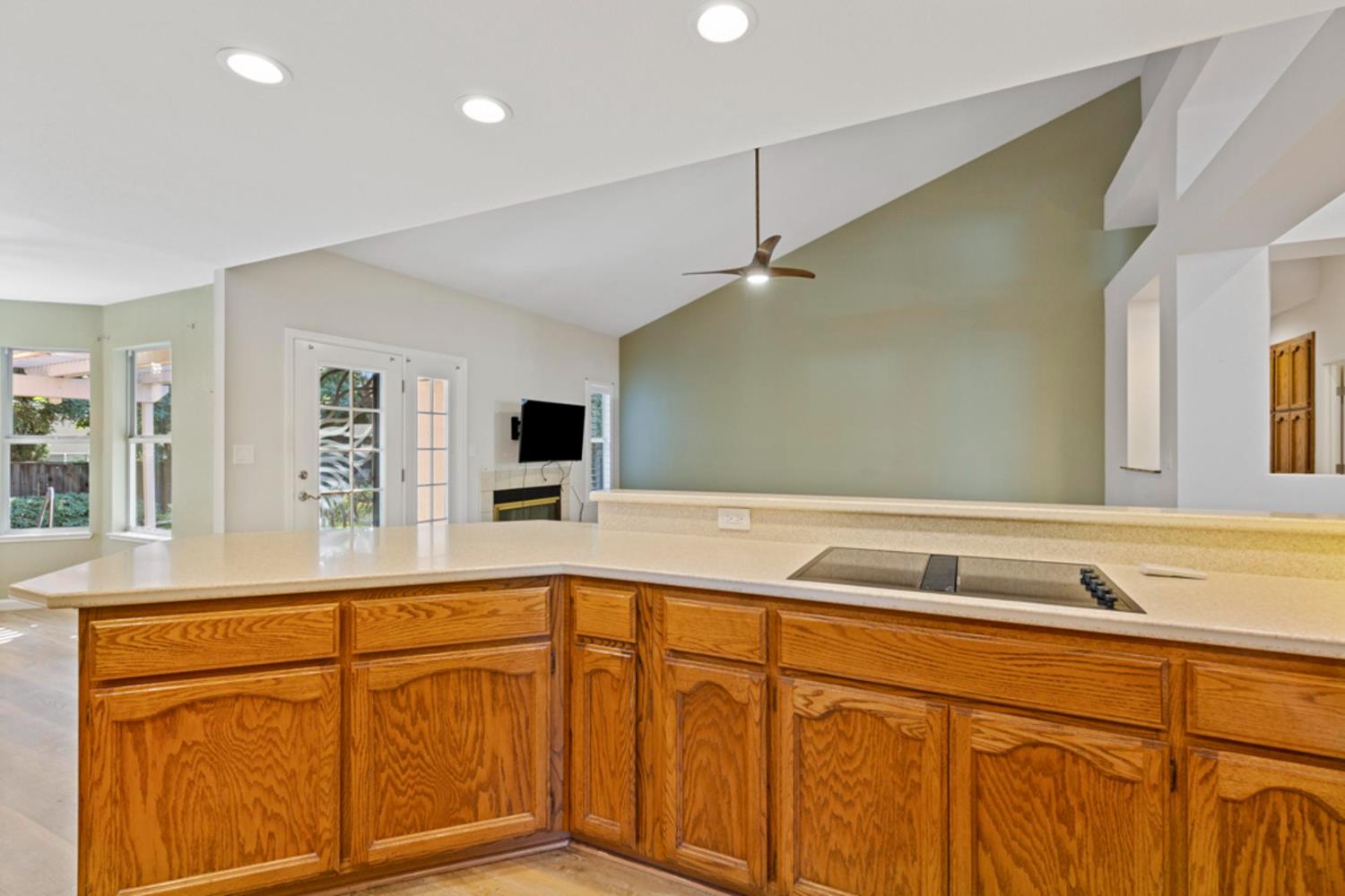 Kitchen peninsula with a cooktop and honey-colored wood cabinets, opening to a vaulted living area.