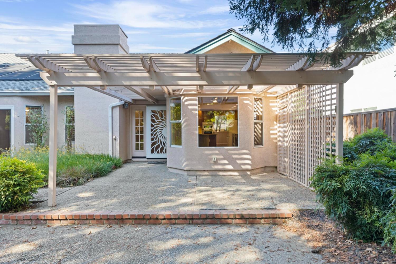 Home’s rear entrance beneath a broad pergola, with a decorative cutout security door and garden beds.