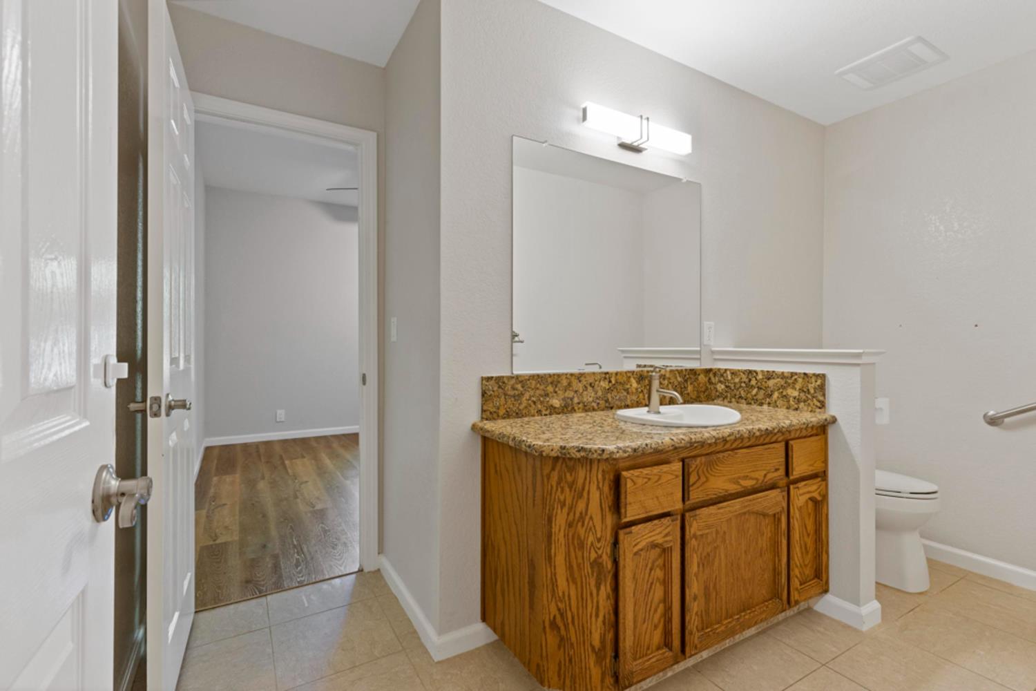 Bathroom with a wood vanity, stone countertop, large mirror, and toilet beside an open doorway.