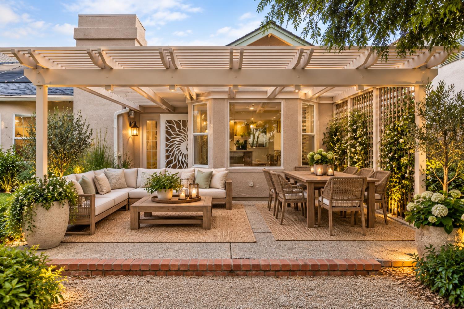 Backyard patio with a white pergola, cushioned sectional, outdoor dining set, and warm landscape lighting.