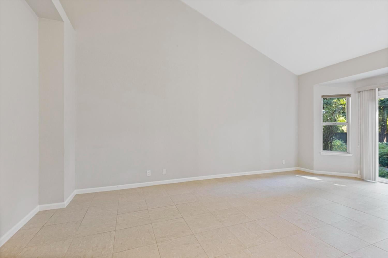 Vacant living room with vaulted ceiling, tiled floor, and sliding glass door beside a window.