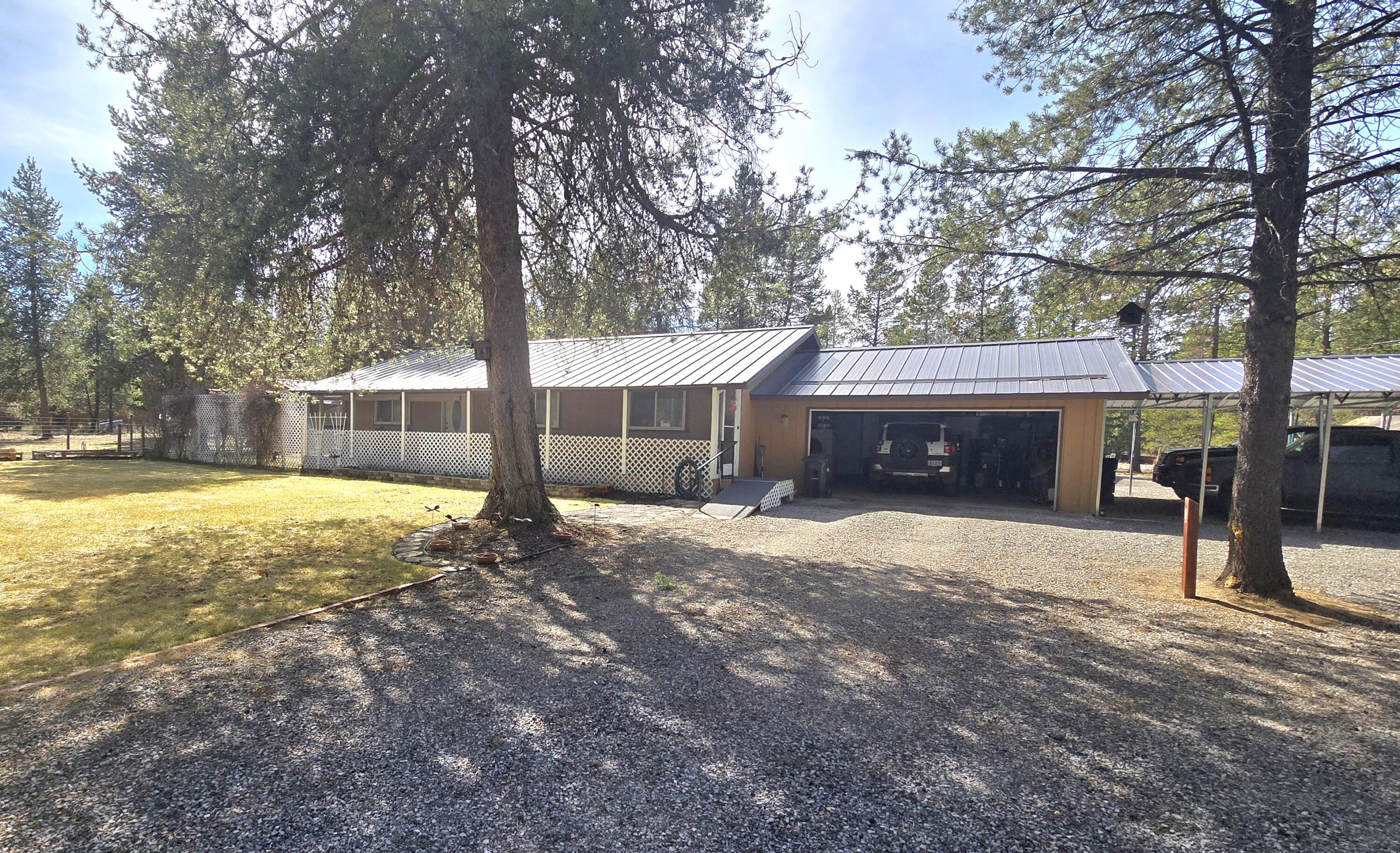 Bend office exterior with a metal roof, covered porch, open garage, and gravel driveway among tall trees.