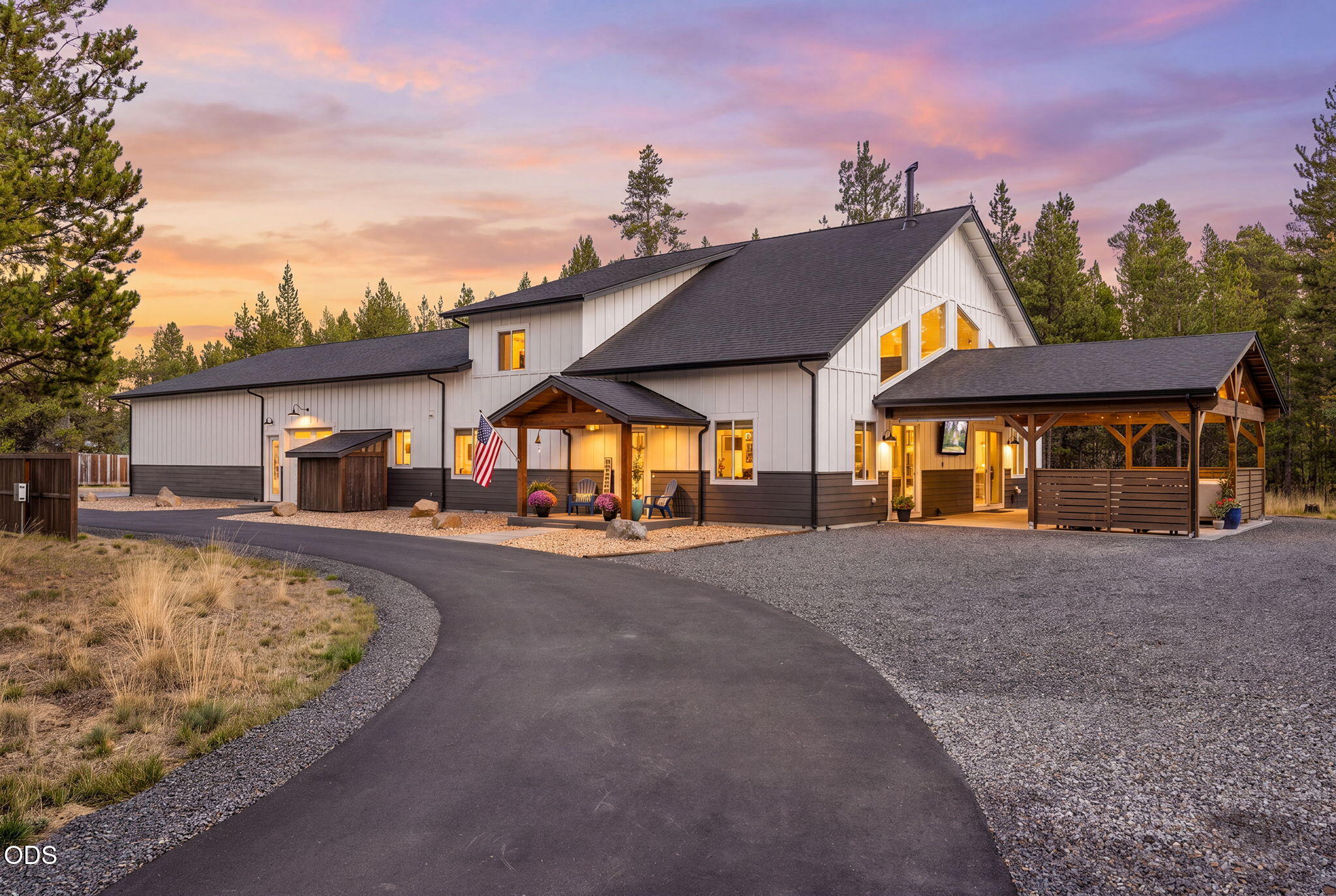 White modern farmhouse among pine trees at dusk, with a covered porch, open carport, and curved driveway.