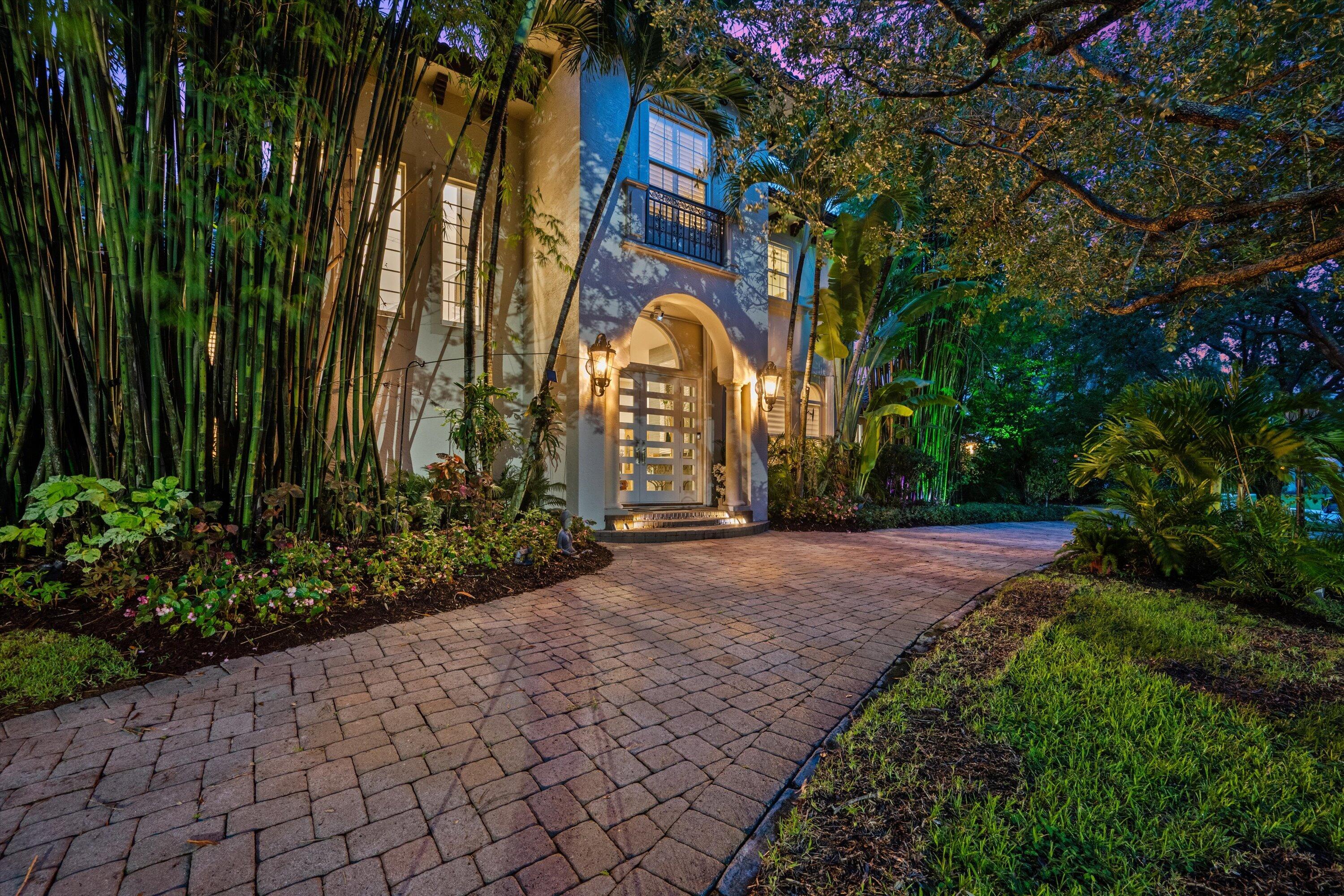 Home entrance with an arched doorway, brick driveway, and lush tropical landscaping at dusk.