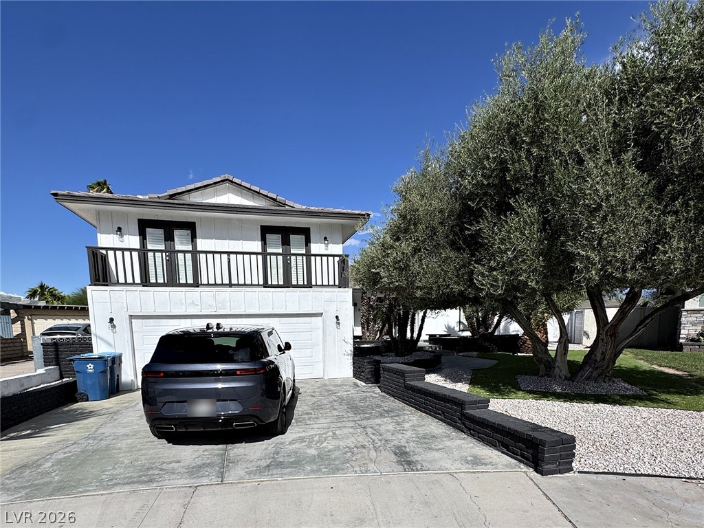 White two-story house with an upper balcony and garage, viewed from the driveway beneath mature trees.