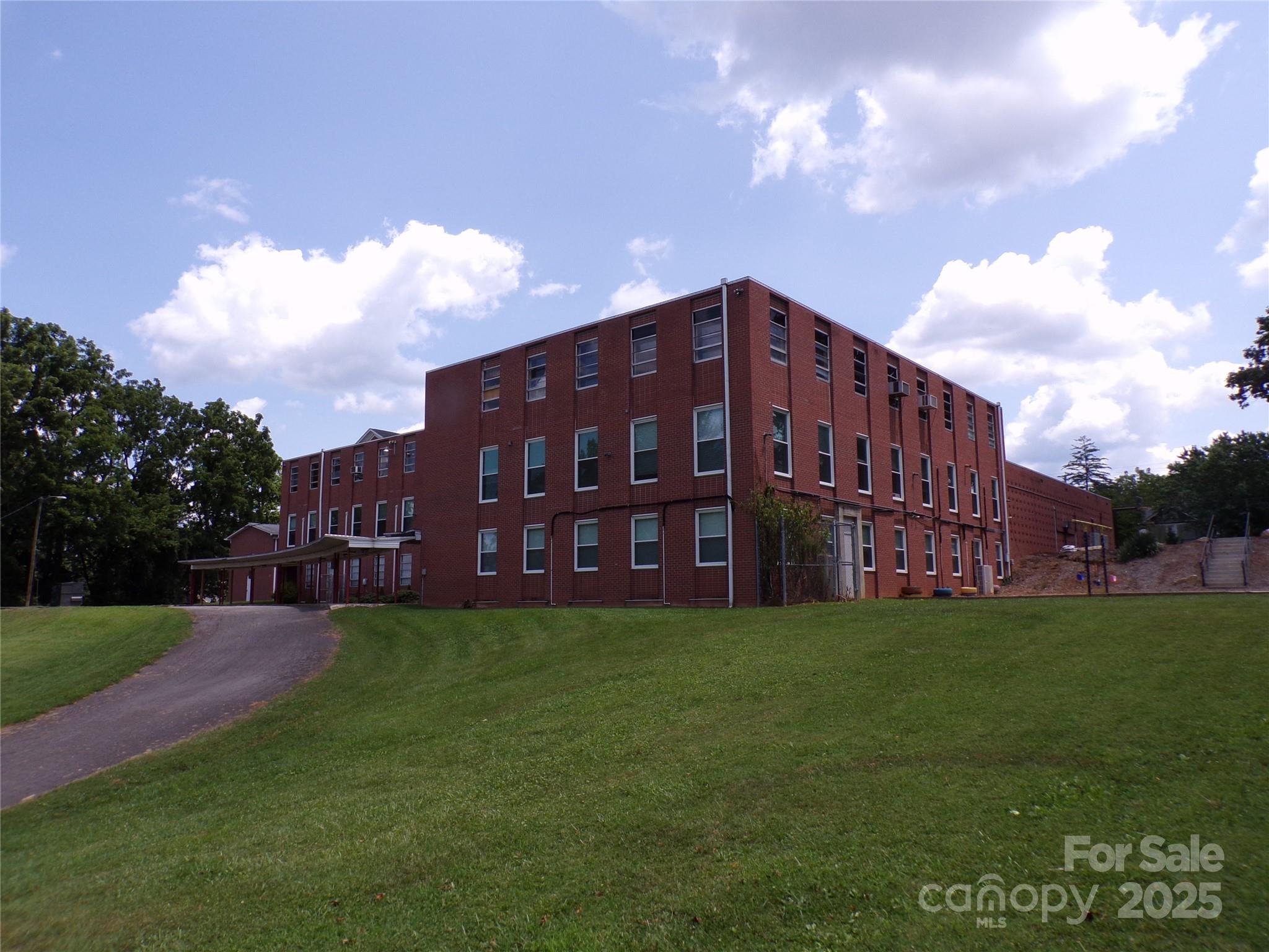 Red brick building with rows of windows on a grassy slope, with a covered entrance and paved drive.