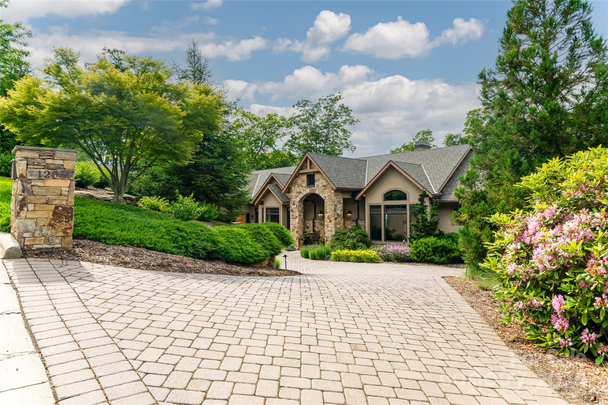 Gabled stone-and-stucco house behind a broad paver driveway, framed by mature trees and flowering shrubs.