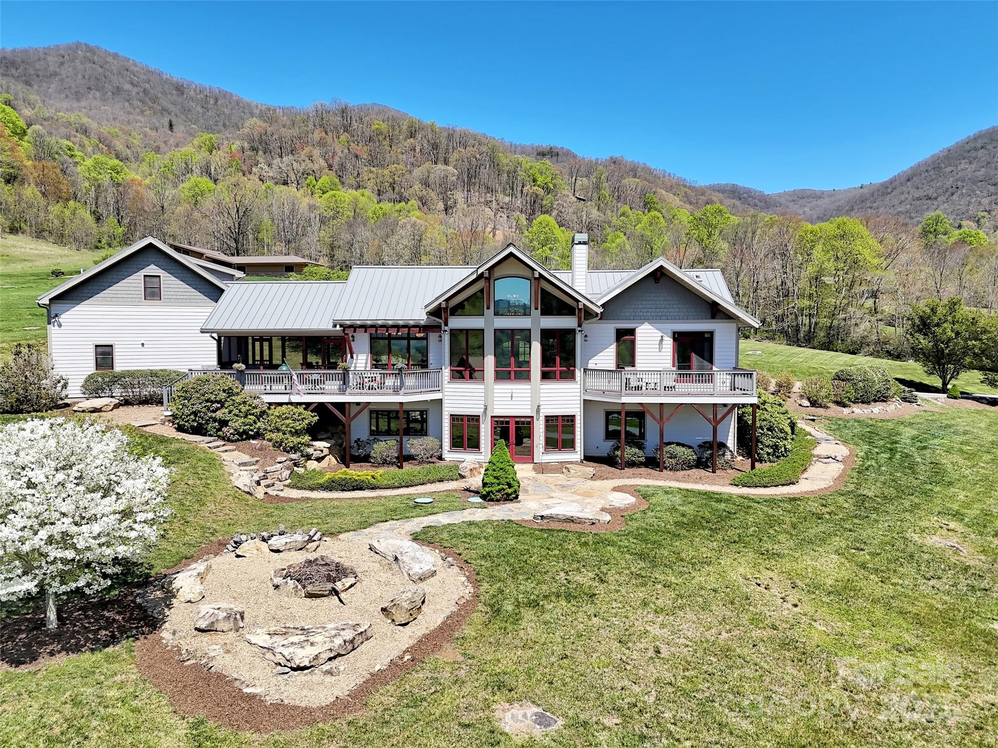 White two-story house with red-trimmed windows, stone gardens, and wooded hills behind it.