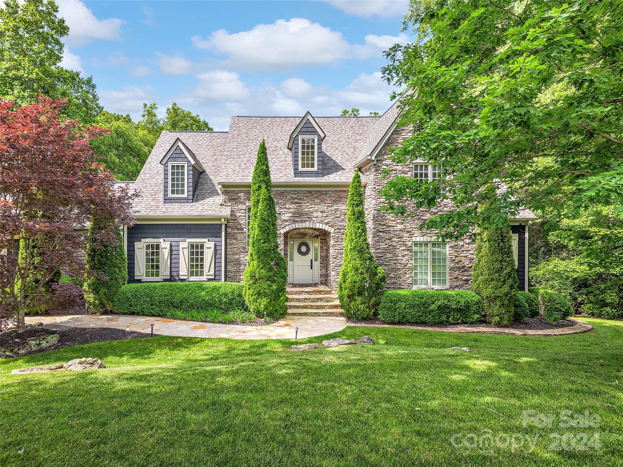 Stone-front home with dormer windows, tall evergreens, clipped hedges, and a broad lawn.