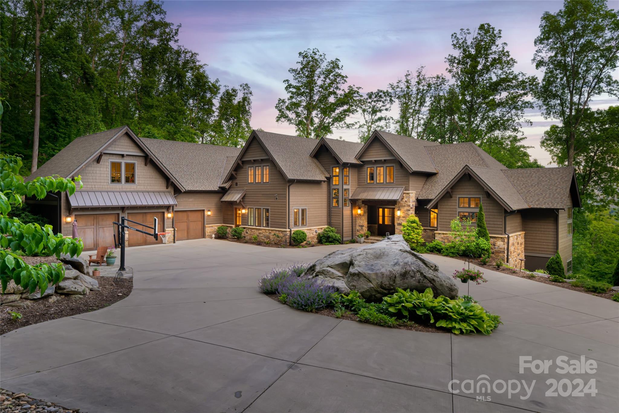Large gabled home with stone accents, a broad driveway, basketball hoop, and wooded surroundings at dusk.