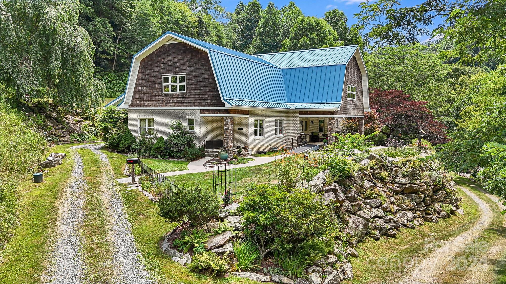 House with a blue metal roof and white brick exterior, surrounded by wooded landscaping and stone walls.