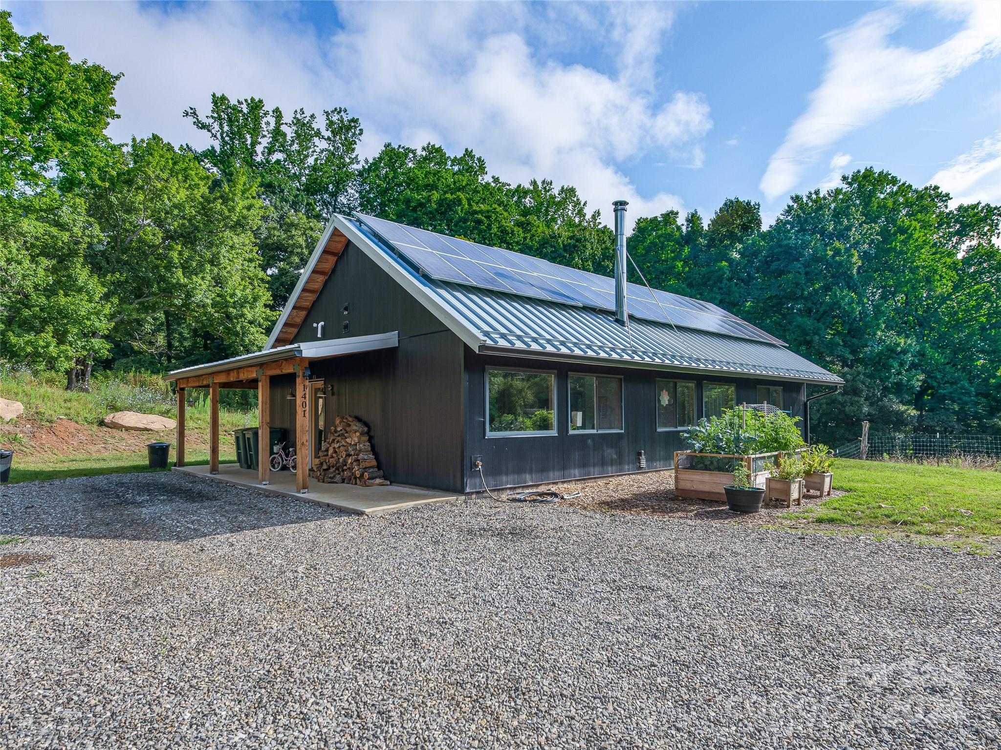Dark modern home with rooftop solar panels, a covered porch, and raised garden beds in a wooded setting.
