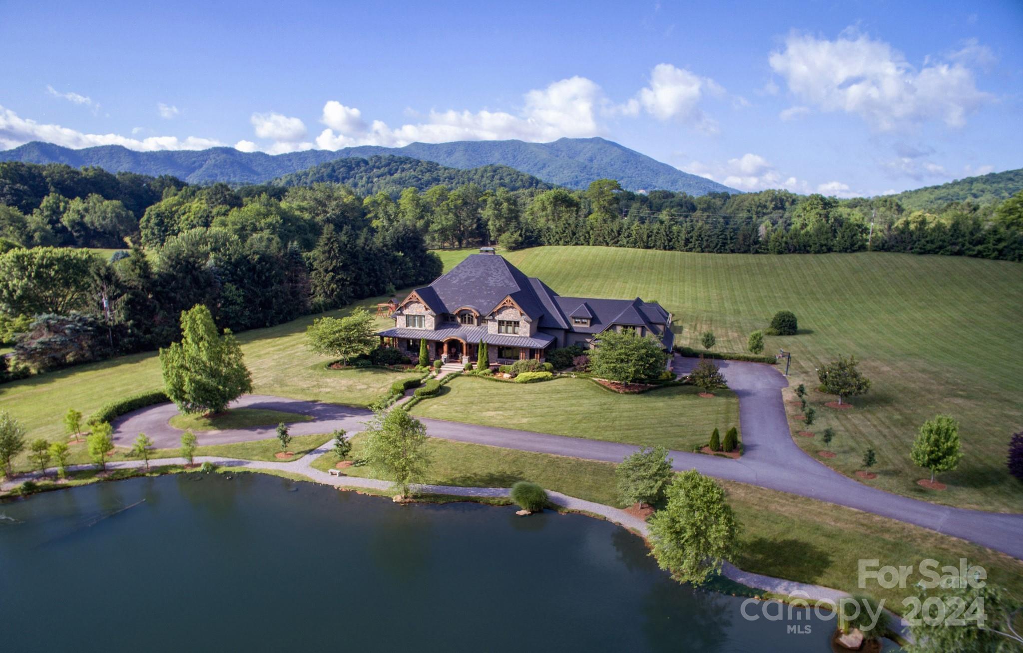 Aerial view of a stone-and-wood home beside a pond, with landscaped grounds and mountains beyond.
