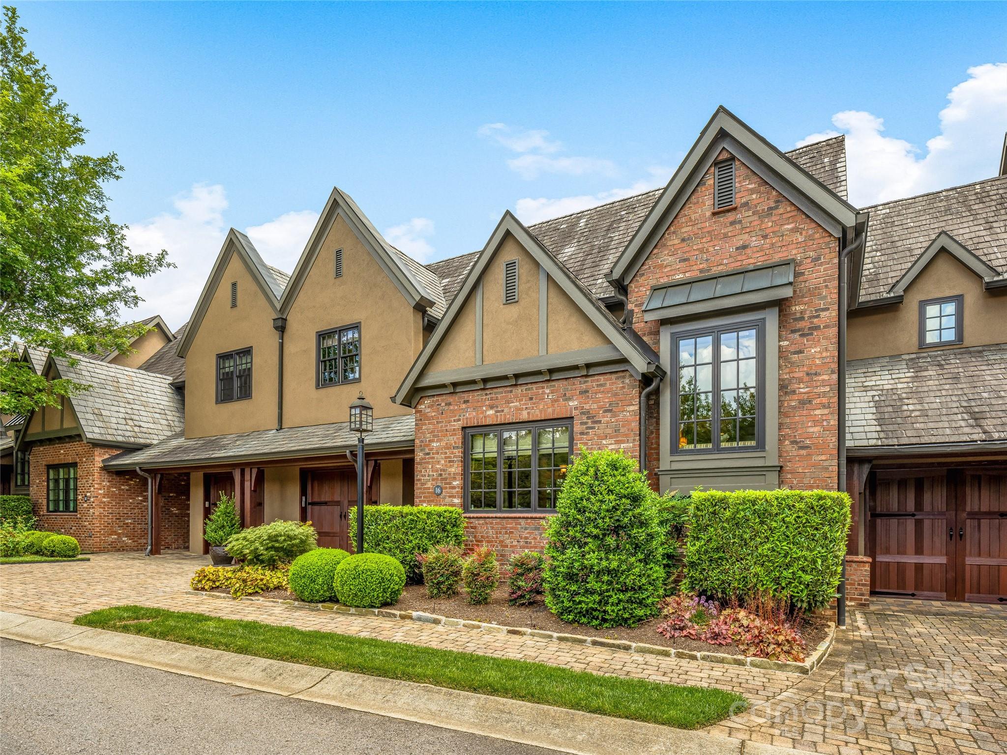 Attached brick and stucco home with steep gables, landscaped shrubs, and wooden garage doors.