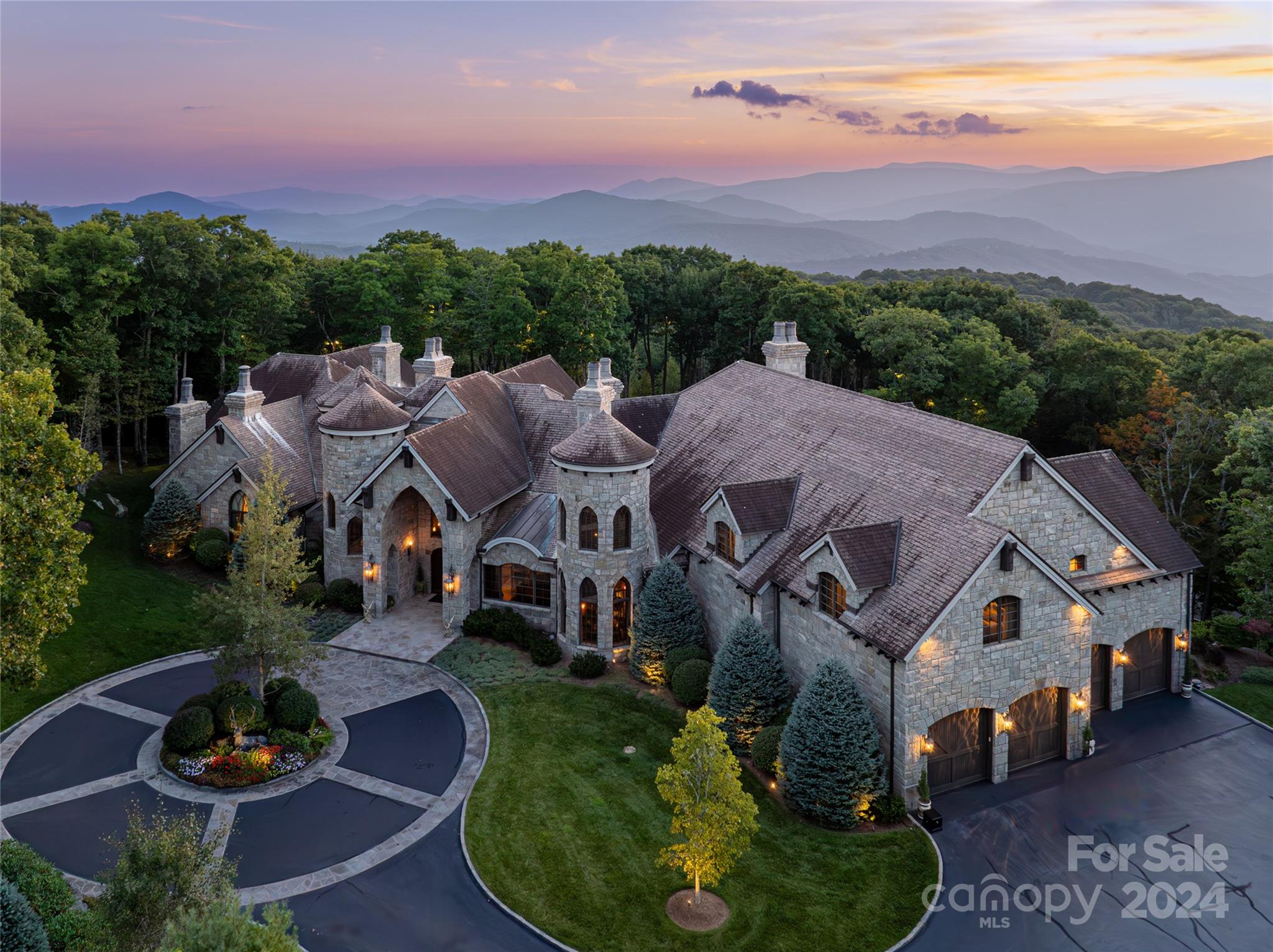 Aerial view of a stone mountain home at dusk, with wooded grounds and layered mountain ridges.