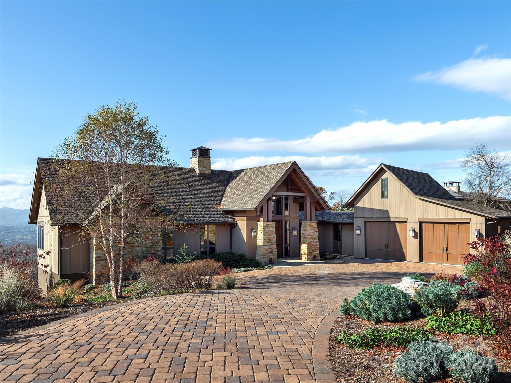 Mountain-style home with stone columns, timber gables, and a broad paver driveway, with distant mountains.