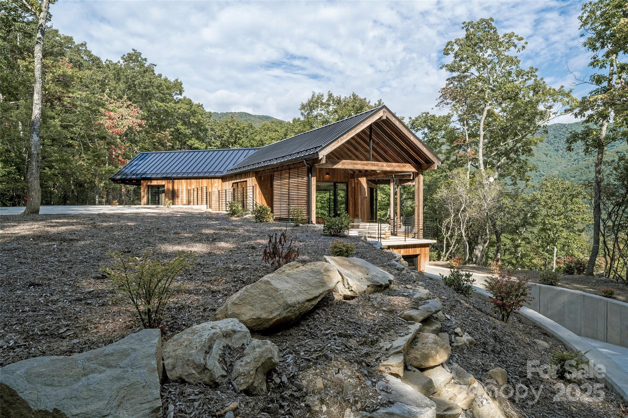 Modern wood-sided home with a metal roof and covered porch among trees overlooking wooded mountains.