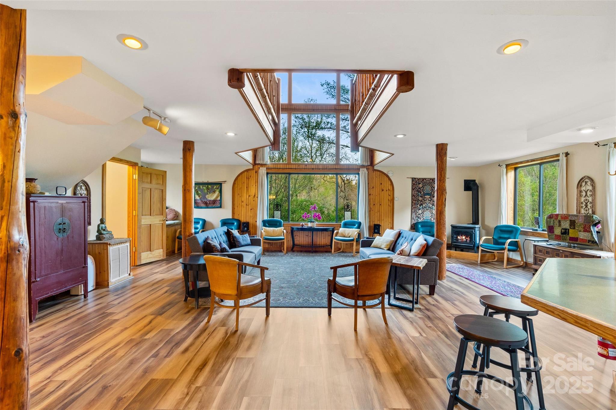 Furnished living room with vaulted ceilings, timber posts, and a tall window overlooking trees.