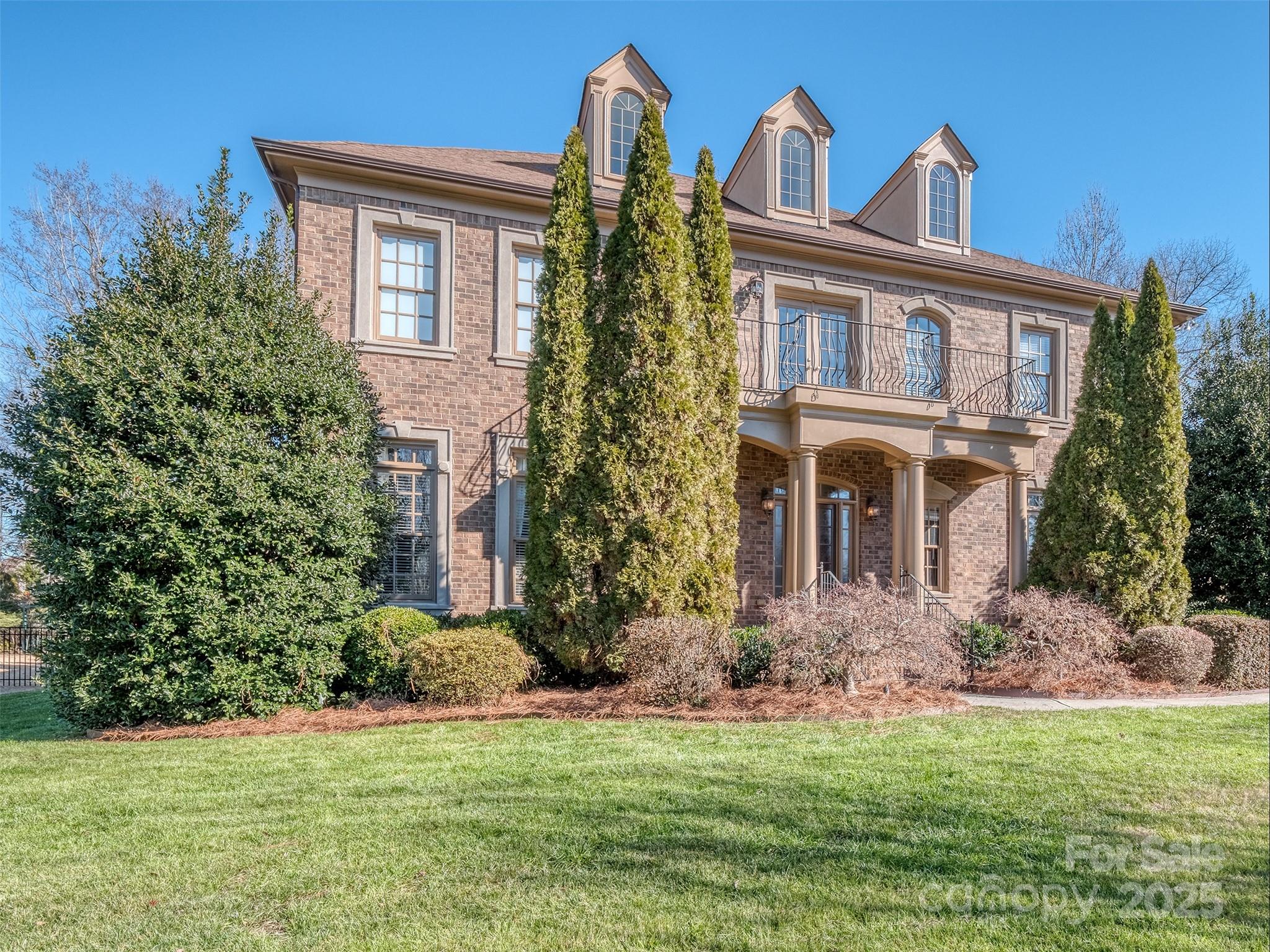 Two-story brick home with three dormers, balconies, tall evergreens, and a landscaped front lawn.