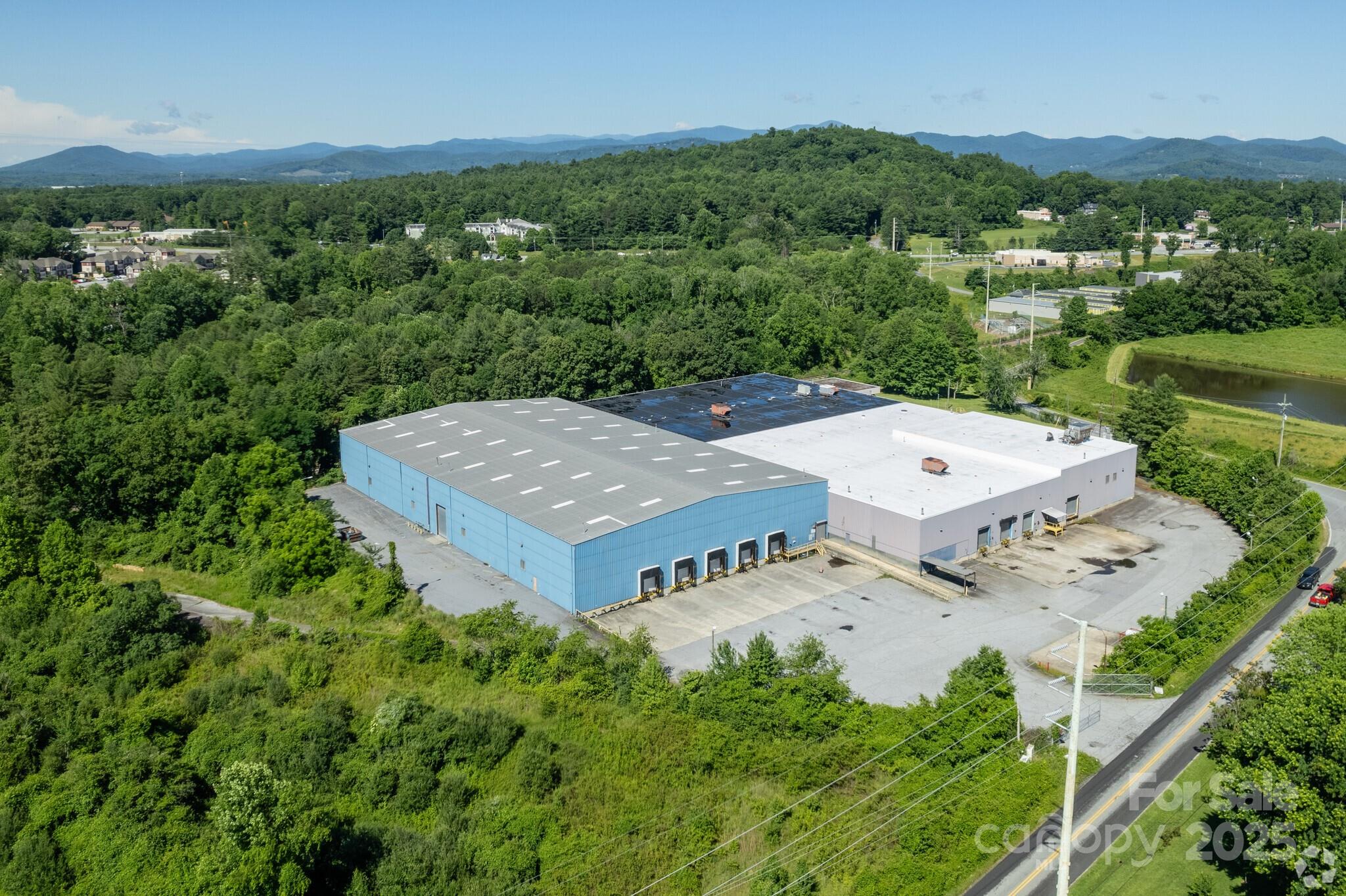 Aerial view of a blue-and-white warehouse with loading docks, wooded hills, and mountains beyond.