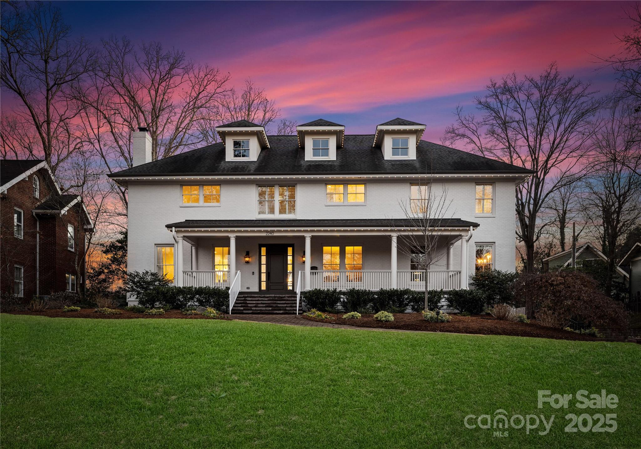 White two-story home with a wide covered porch and glowing windows beneath a colorful dusk sky.