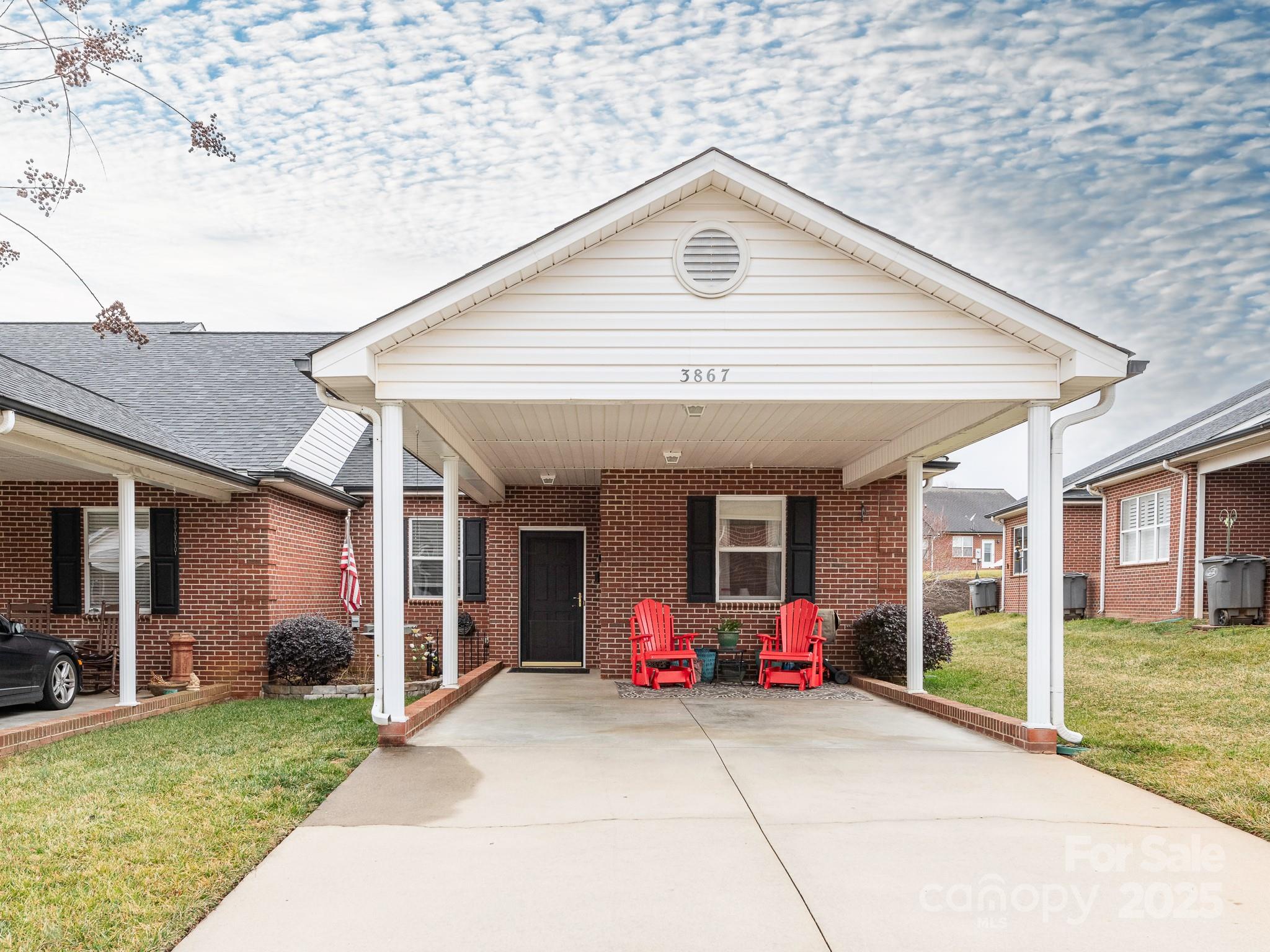 Red-brick home with a broad covered carport, concrete driveway, and two red chairs by the entrance.