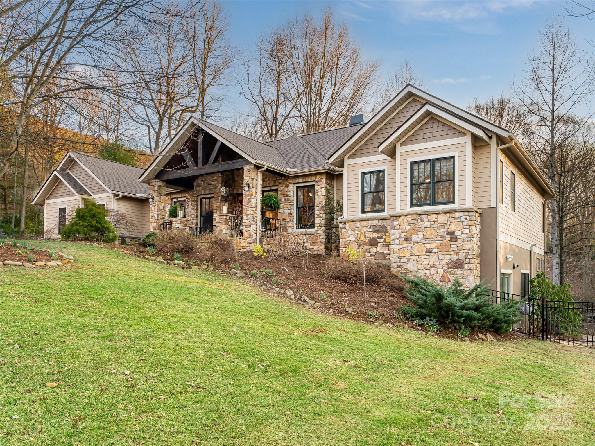 Home with a stone façade and covered entrance on a sloping lawn among leafless trees.