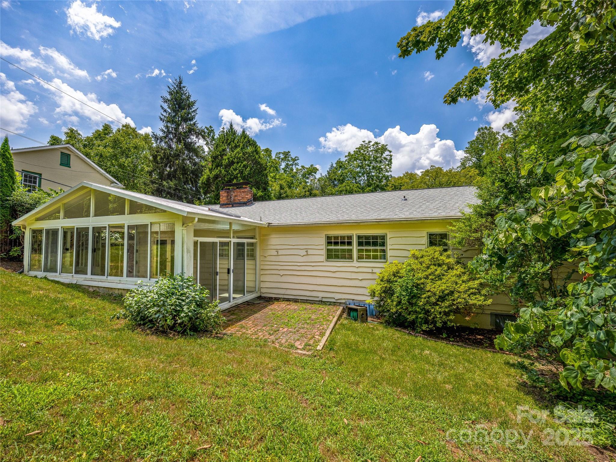 Rear of a light-colored house with a glass sunroom, brick patio, and leafy yard.